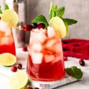 A vibrant cranberry mocktail with ice, garnished with cranberries, mint, and a lime slice, sits elegantly on a marble surface surrounded by scattered cranberries and lime wedges.