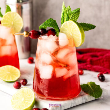 A vibrant cranberry mocktail with ice, garnished with cranberries, mint, and a lime slice, sits elegantly on a marble surface surrounded by scattered cranberries and lime wedges.