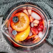 Top view of a glass pitcher brimming with red sangria, filled with ice, fruit slices, and berries, resting on a textured gray surface.