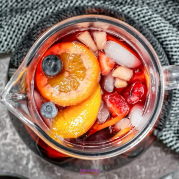 Top view of a glass pitcher brimming with red sangria, filled with ice, fruit slices, and berries, resting on a textured gray surface.