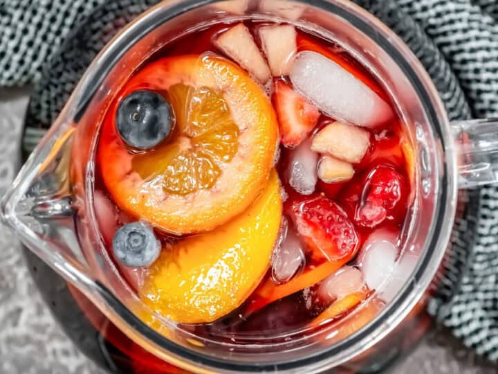 Top view of a glass pitcher brimming with red sangria, filled with ice, fruit slices, and berries, resting on a textured gray surface.