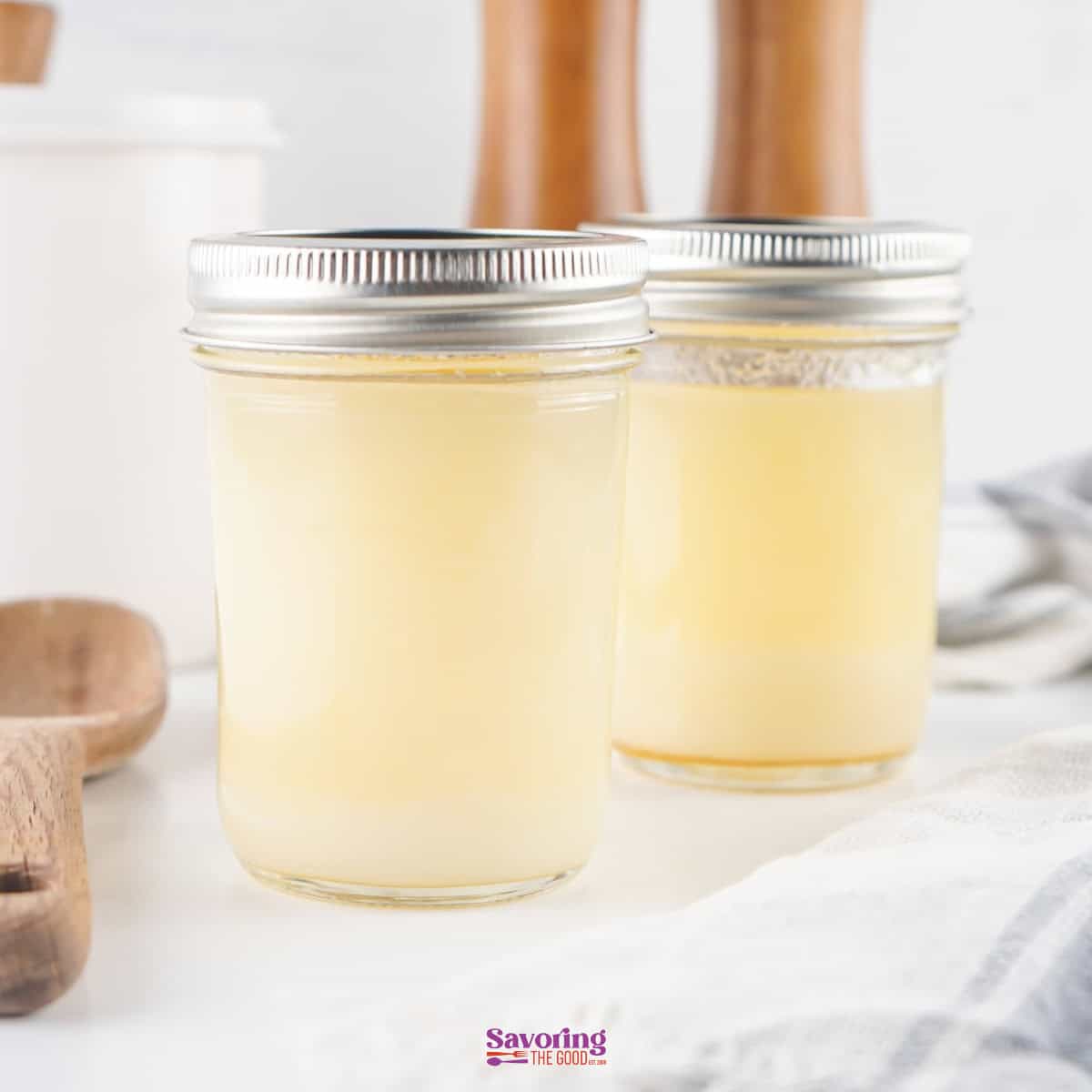Two glass jars filled with a light-colored liquid, resembling melted beef tallow, sit on a white surface. Nearby, wooden kitchen utensils and a striped cloth complete the simple yet elegant arrangement.