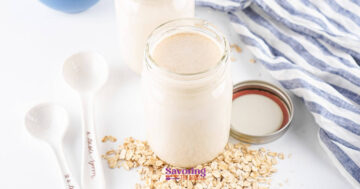A jar of homemade oat milk surrounded by scattered oats, a striped cloth, and measuring spoons on a white surface.
