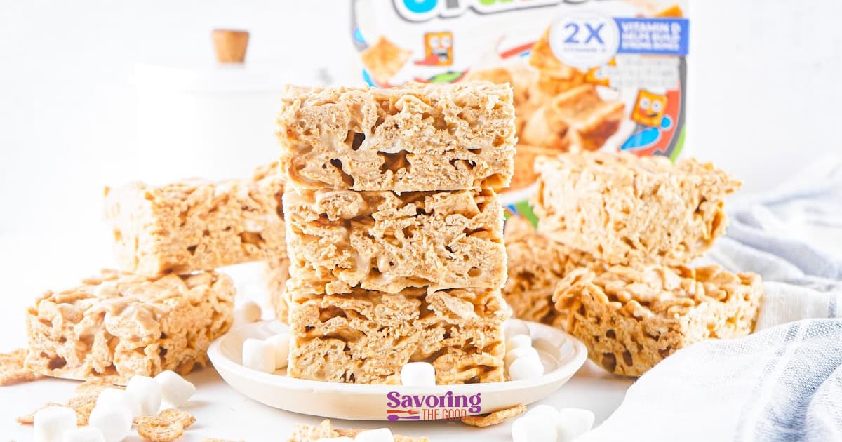 Cinnamon Toast Crunch bars are stacked on a plate, surrounded by more treats, mini marshmallows, and a cereal box in the background.