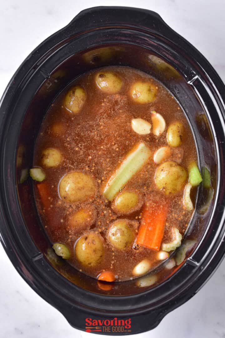 A Crock Pot simmers a hearty stew with potatoes, carrots, celery, and garlic cloves in a rich brown broth. Perfectly flavored corned beef adds depth, while tender cabbage completes this comforting dish.
