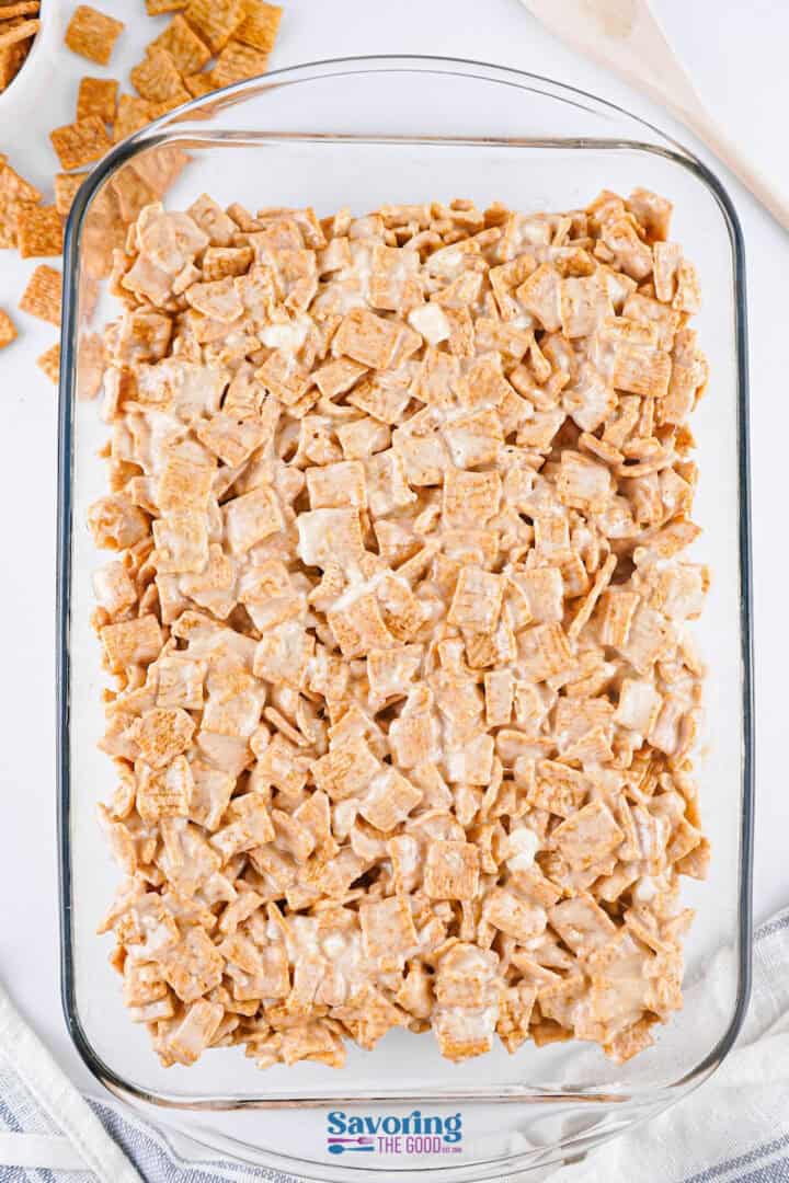 A glass dish brimming with Cinnamon Toast Crunch Bars, surrounded by a spoon and colorful cereal pieces on the side.
