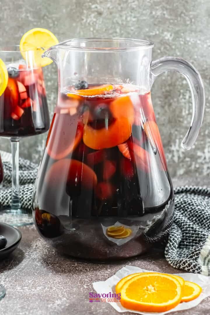 A glass pitcher filled with dark red sangria, containing slices of orange and blueberries. A glass of sangria is in the background, and orange slices are placed nearby.