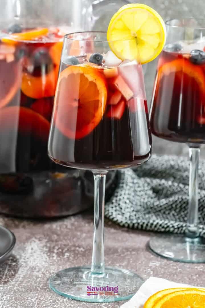 A glass of dark red sangria with orange and lemon slices, garnished with blueberries, set on a table with more sangria and fruit in the background.
