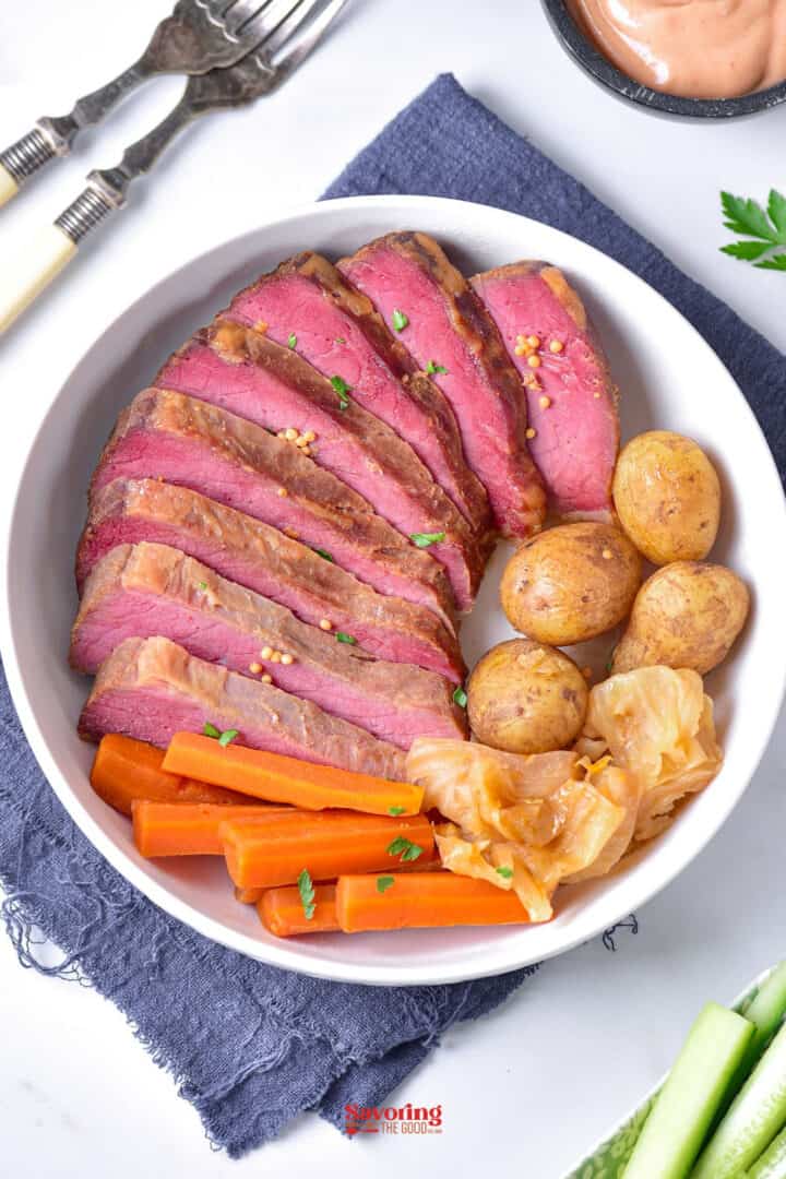 Sliced roast beef with boiled potatoes, carrots, and cabbage in a white bowl, set on a blue cloth napkin with forks and a sauce on the side.