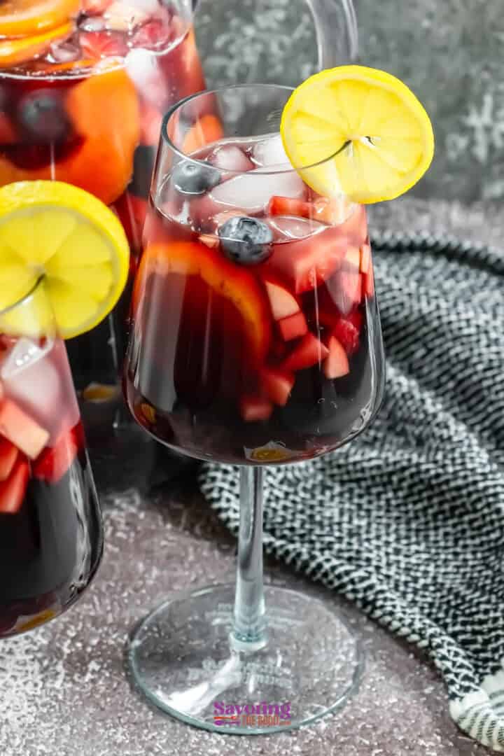 A glass of sangria with ice, apple cubes, orange slices, blueberries, and a lemon slice on the rim.