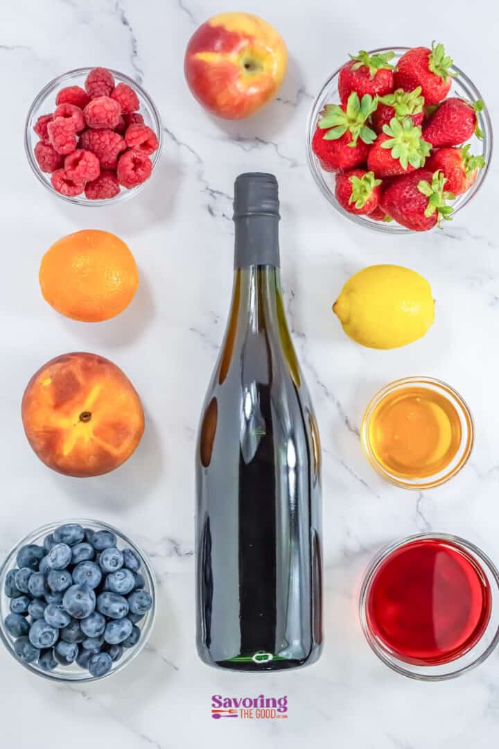 A bottle of wine is surrounded by bowls of raspberries, strawberries, blueberries, an orange, a lemon, two peaches, a bowl of honey, and a glass of red liquid on a white marble surface.