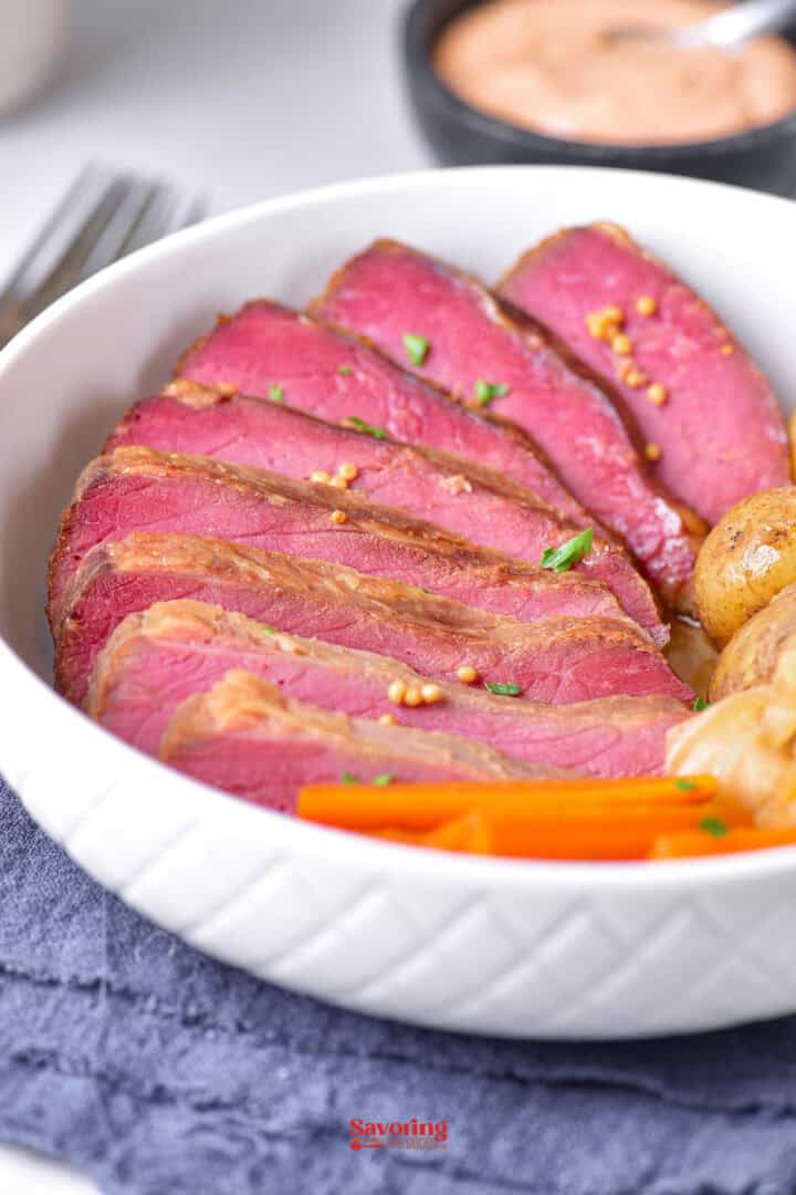 Slices of cooked corned beef with carrots and potatoes in a white bowl, placed on a blue cloth.