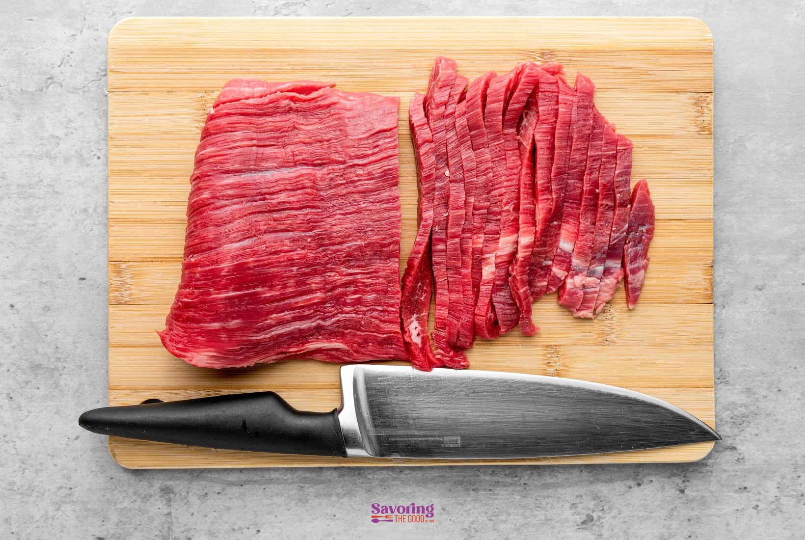 A cutting board with a piece of flank steak, half whole and half sliced into thin strips, beside a large kitchen knife.