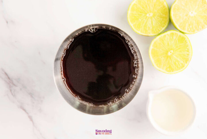 A metal cup filled with dark liquid next to halved limes and a small white pitcher on a marble surface.