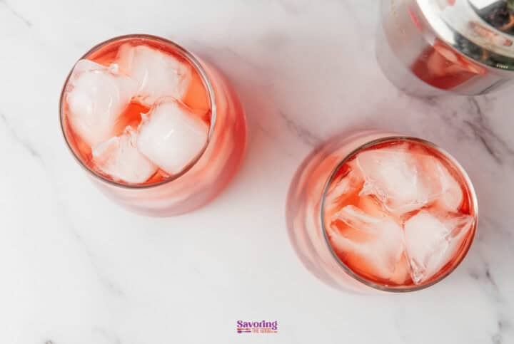 Two glasses of a pink cocktail with ice cubes on a marble surface, next to a metal shaker.