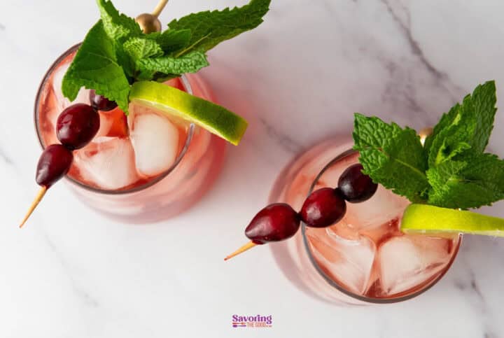 Two glasses of refreshing cranberry mocktails, garnished with lime slices, mint leaves, and skewered cherries, rest enticingly on a marble surface.