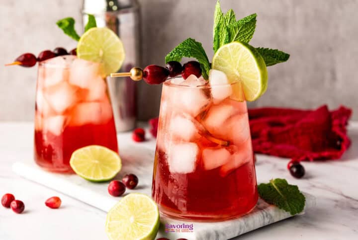 Two refreshing cranberry mocktails in glasses, adorned with lime slices, mint sprigs, and cranberries on skewers, elegantly set on a marble surface.