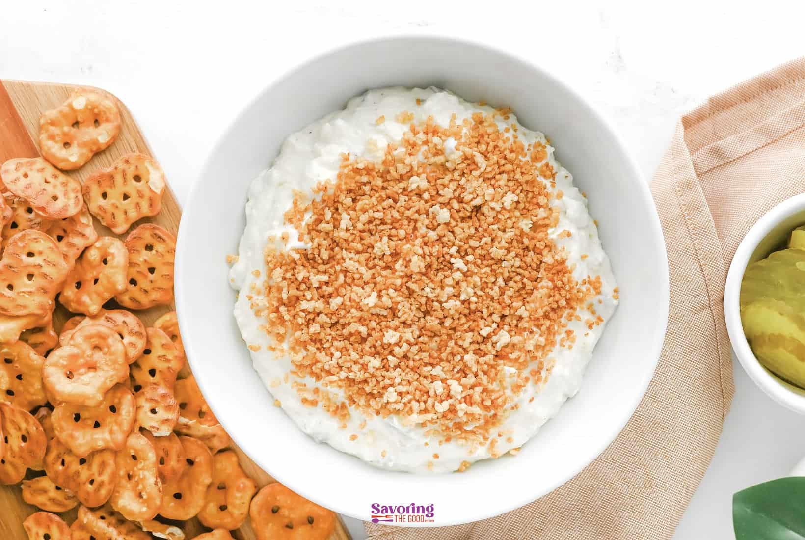 A bowl of fried dill pickle dip topped with breadcrumbs, surrounded by pretzel crisps and a bowl of pickles.