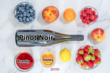 A bottle labeled "Pinot Noir" is surrounded by ingredients perfect for a red sangria: blueberries, orange, raspberries, apple, strawberries, peach, lemon, a bowl of brandy, and a bowl of honey on a marble surface.