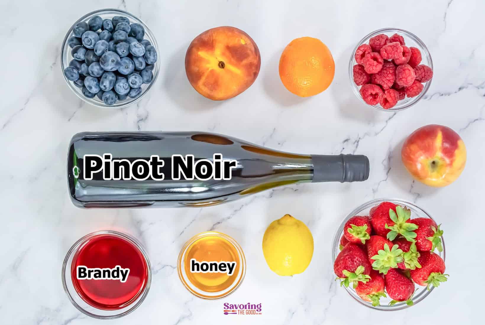 A bottle labeled "Pinot Noir" is surrounded by ingredients perfect for a red sangria: blueberries, orange, raspberries, apple, strawberries, peach, lemon, a bowl of brandy, and a bowl of honey on a marble surface.