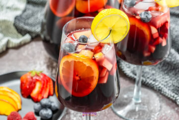 Glasses of red sangria with fruit slices and berries, garnished with lemon, sit on a table alongside a plate of fresh fruit.