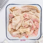 Raw beef fat pieces in a glass dish next to a chef's knife and striped towel on a white surface.