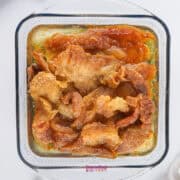 A glass dish with crispy beef fat pieces, glistening in a 9x9 pan in beef tallow, rests on a white surface, partially covered by a white cloth.