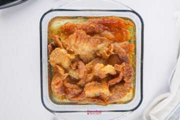 A glass dish with crispy beef fat pieces, glistening in a 9x9 pan in beef tallow, rests on a white surface, partially covered by a white cloth.