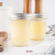 Two sealed mason jars filled with homemade beef tallow are on a white surface, next to a wooden spoon and a striped cloth. A white container and wooden mills are in the background.