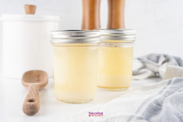 Two sealed mason jars filled with homemade beef tallow are on a white surface, next to a wooden spoon and a striped cloth. A white container and wooden mills are in the background.