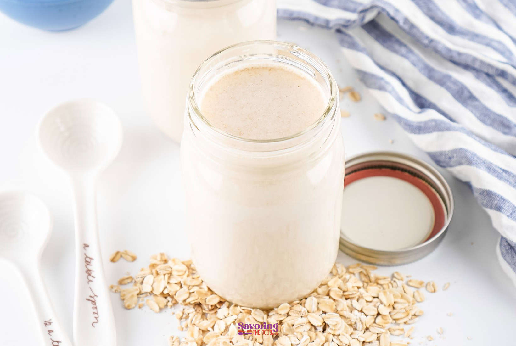 A jar of oat milk surrounded by loose oats, with a striped cloth and measuring spoons nearby. An open lid is placed alongside the jar.