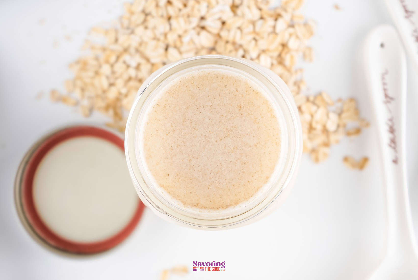 A jar is seen from above, surrounded by scattered oats and a hint of oat milk, with a lid and spoon nearby.