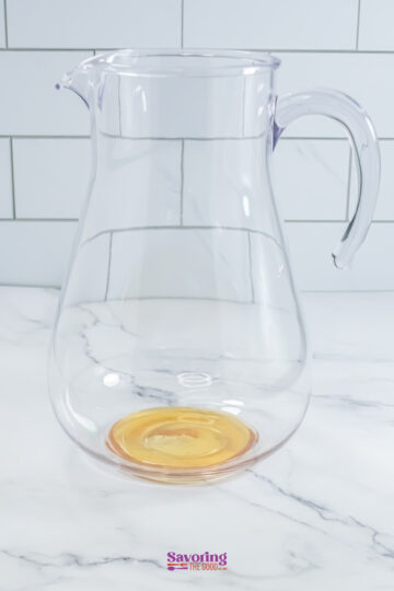 A clear glass pitcher with a small amount of liquid at the bottom, placed on a marble surface with a tiled background.
