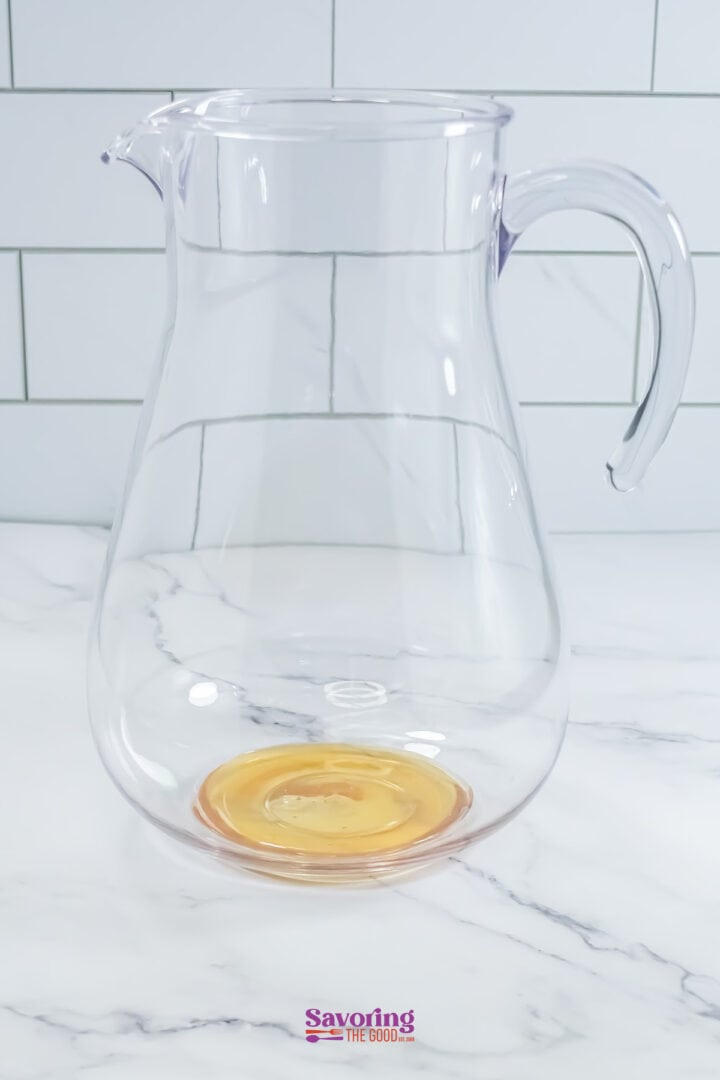 A clear glass pitcher with a small amount of liquid at the bottom, placed on a marble surface with a tiled background.