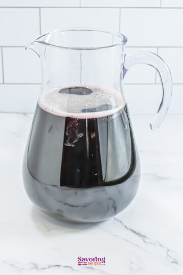 A clear pitcher filled with dark red sangria sits on a marble surface, against a white tiled background.