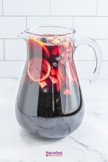 A glass pitcher filled with red sangria, featuring visible slices of citrus, apple chunks, and berries.