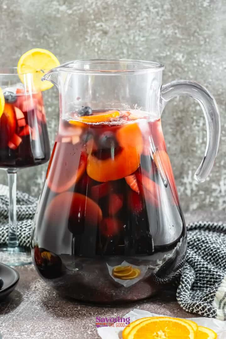 A clear pitcher brimming with vibrant red sangria showcases slices of oranges, apples, and berries. In the background, a glass of this delightful red sangria is garnished with a lemon slice for an extra touch of zest.