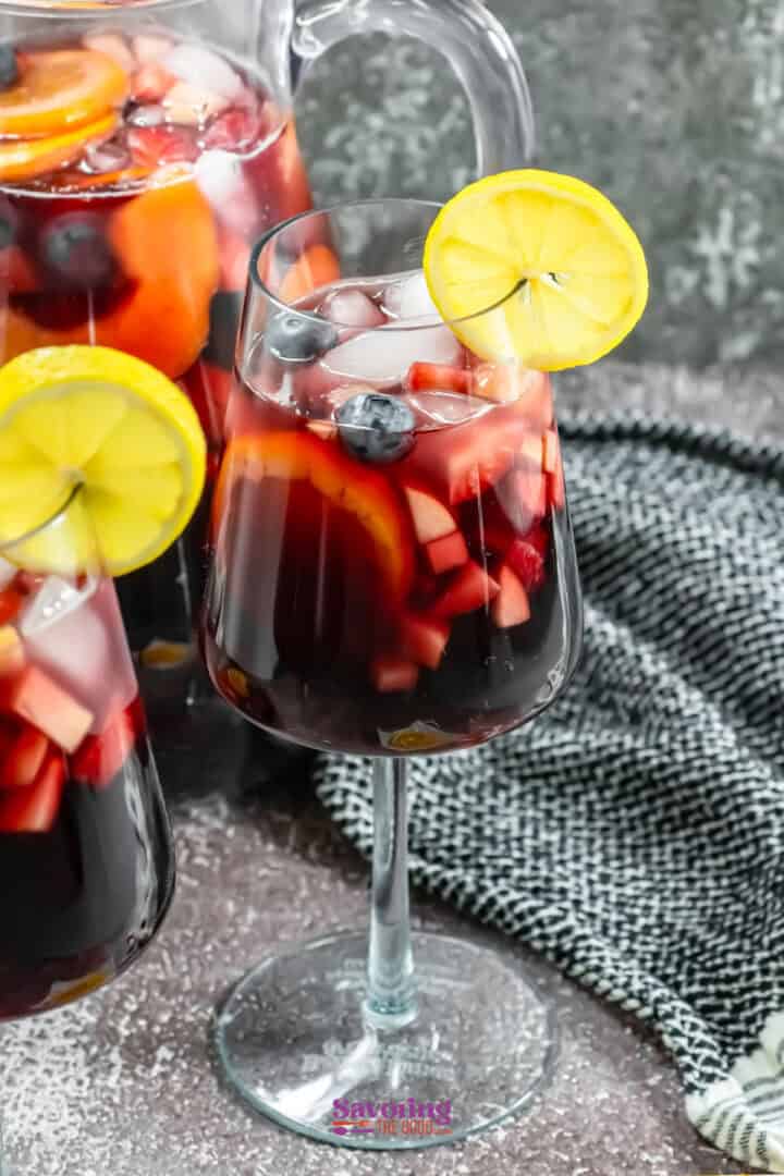 Glass of red sangria with fruit slices and a lemon garnish, next to a pitcher on a textured surface with a black and white cloth.