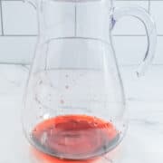 A clear glass pitcher containing a small amount of red liquid at the bottom, placed on a marble surface with a tiled wall in the background.
