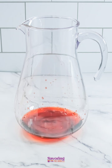 A clear glass pitcher containing a small amount of red liquid at the bottom, placed on a marble surface with a tiled wall in the background.