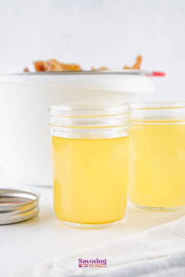 Two jars of clear beef tallow, sit on a white surface, with a pot in the background.
