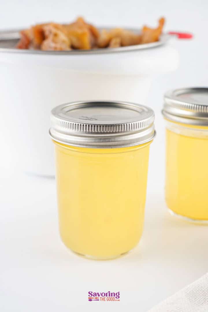 Two clear jars filled with beef tallow that is still liquid, capped with silver lids, containing rendered beef tallow. Blurred in the background is a white bowl with small pieces of food.