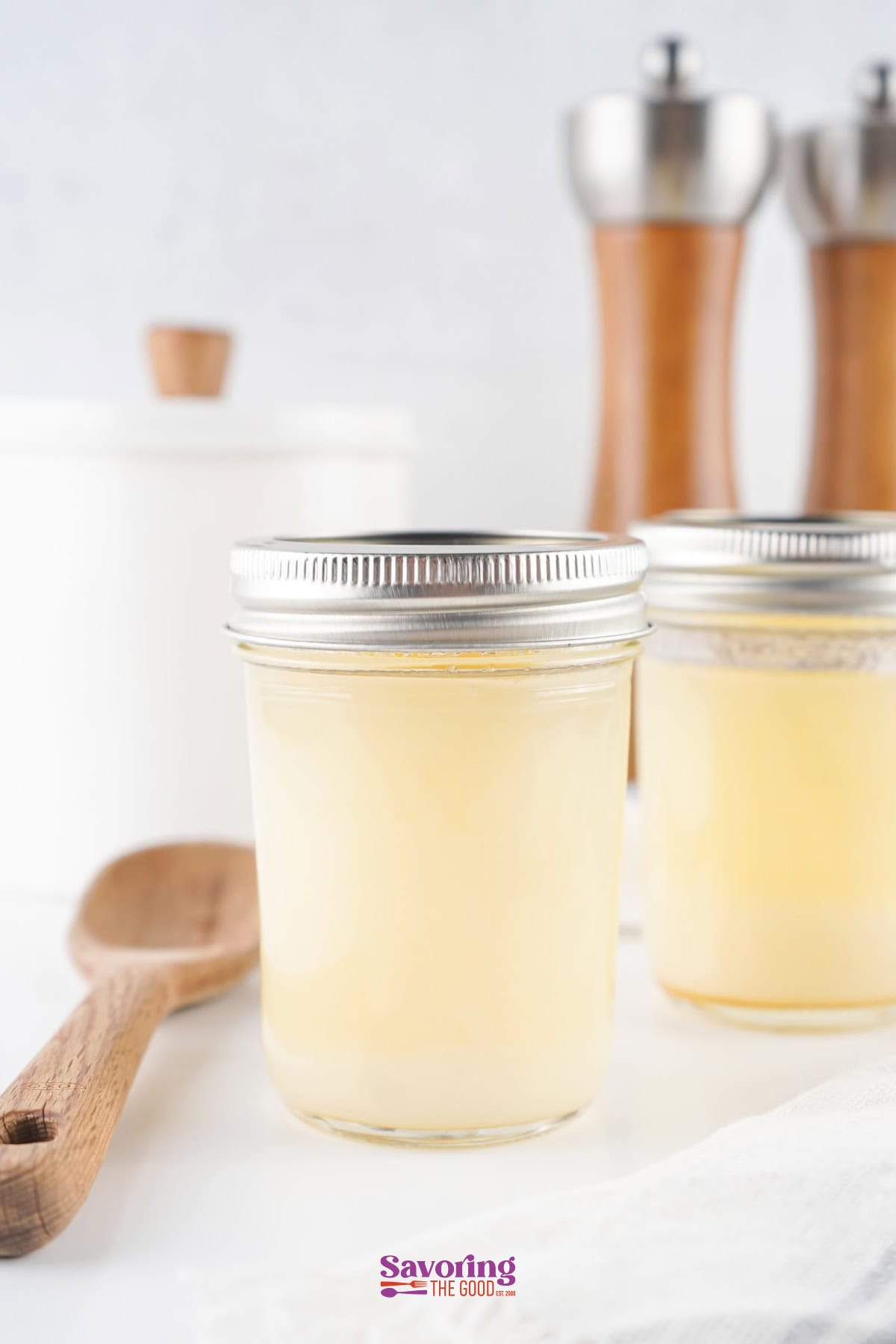 Two glass jars filled with solid beef tallow sit on a white surface, hinting at the rich, smooth essence of beef tallow within. A wooden spoon and pepper mills are artistically blurred in the background, adding a rustic charm to the scene.
