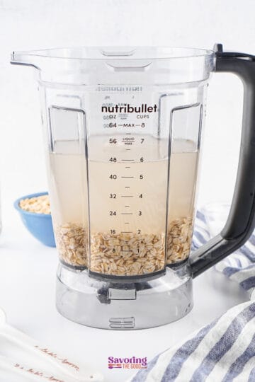 A blender pitcher filled with water and oats, placed on a countertop with a striped cloth nearby.