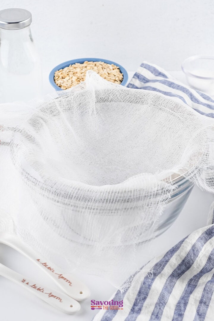 Strainer with cheesecloth over a bowl, next to a bowl of oats, two decorated spoons, a striped cloth, and a bottle of milk on a white surface.