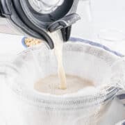 A machine pours liquid through a cheesecloth-lined strainer into a bowl, part of a kitchen setup for making homemade milk.