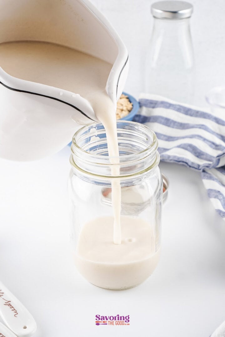 Homemade oatmeal milk cascades from a white jug into a glass jar, accompanied by a striped cloth and a glass bottle in the background.