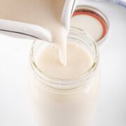 Oatmeal milk cascades from a pitcher into a mason jar, its creamy texture promising nourishment. The lid sits close by, ready to seal in freshness, all against a pristine white background.