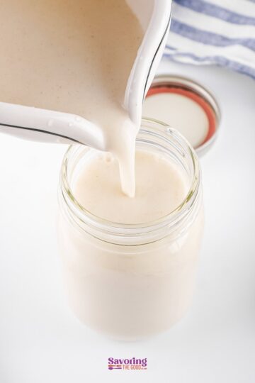 Oatmeal milk cascades from a pitcher into a mason jar, its creamy texture promising nourishment. The lid sits close by, ready to seal in freshness, all against a pristine white background.