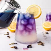 A glass of butterfly pea flower lemonade is being poured, displaying a gradient from blue to purple, garnished with a lemon slice. Lemons and dried flowers are scattered on the white surface.